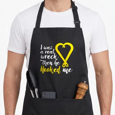 Hooked truck apron - Two yellow hooks form a heart next to stacked white text saying I was a real wreck then he Hooked — shown on a black cotton canvas apron against a white background