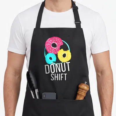 Shift donut apron - Three pink, yellow, cyan donuts with bites taken out above text DONUT SHIFT — shown on a black cotton canvas apron against a white background