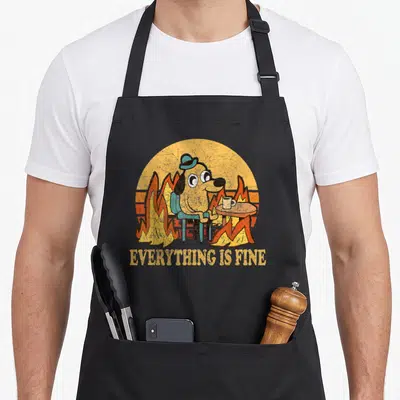 Fine dog apron - Cartoon dog in flames with mug and EVERYTHING IS FINE text — shown on a black cotton canvas apron against a white background