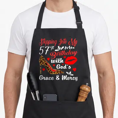 Step birthday apron - Leopard high heel shoe with red lips and 57th Birthday with God's Grace text — shown on a black cotton canvas apron against a white background
