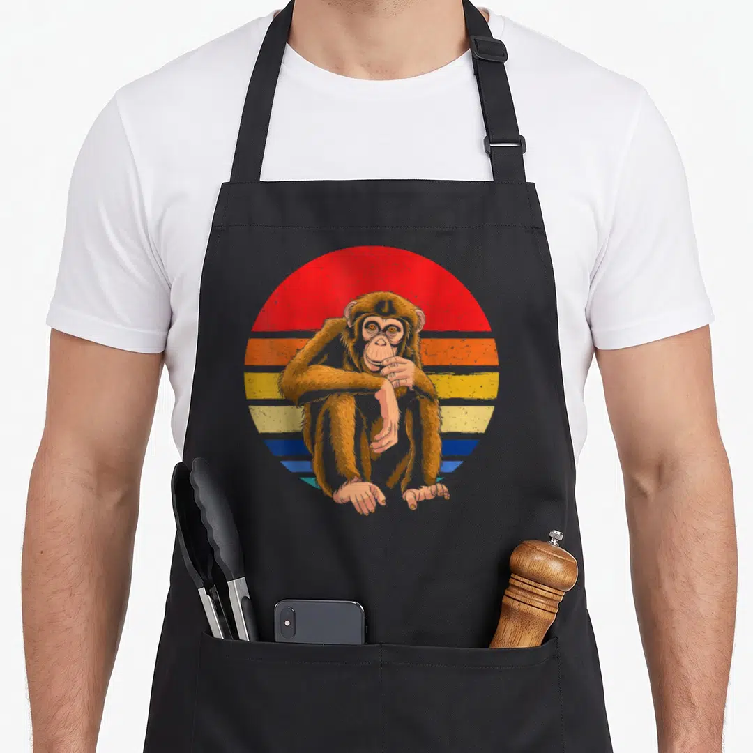 Chimpanzee monkey animal apron - Brown monkey sitting in front of red orange yellow and blue striped circle — shown on a black cotton canvas apron against a white background