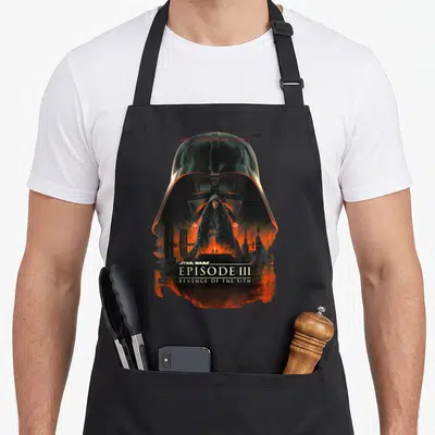 Episode anniversary apron - Darth Vader helmet framing Anakin Skywalker over lava with 20th Anniversary text — shown on a black cotton canvas apron against a white background