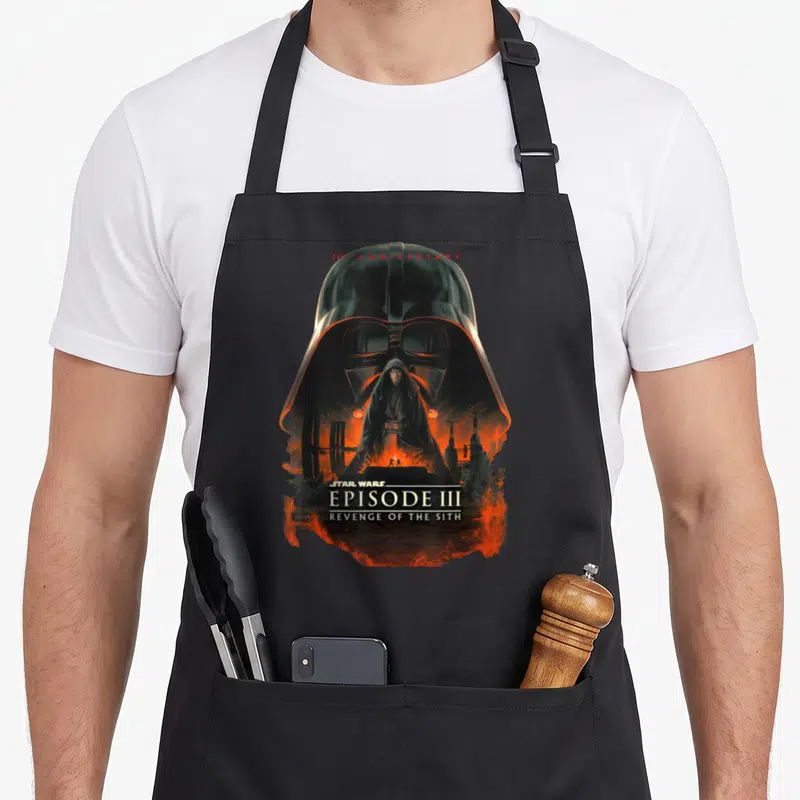 Episode anniversary apron - Darth Vader helmet framing Anakin Skywalker over lava with 20th Anniversary text — shown on a black cotton canvas apron against a white background