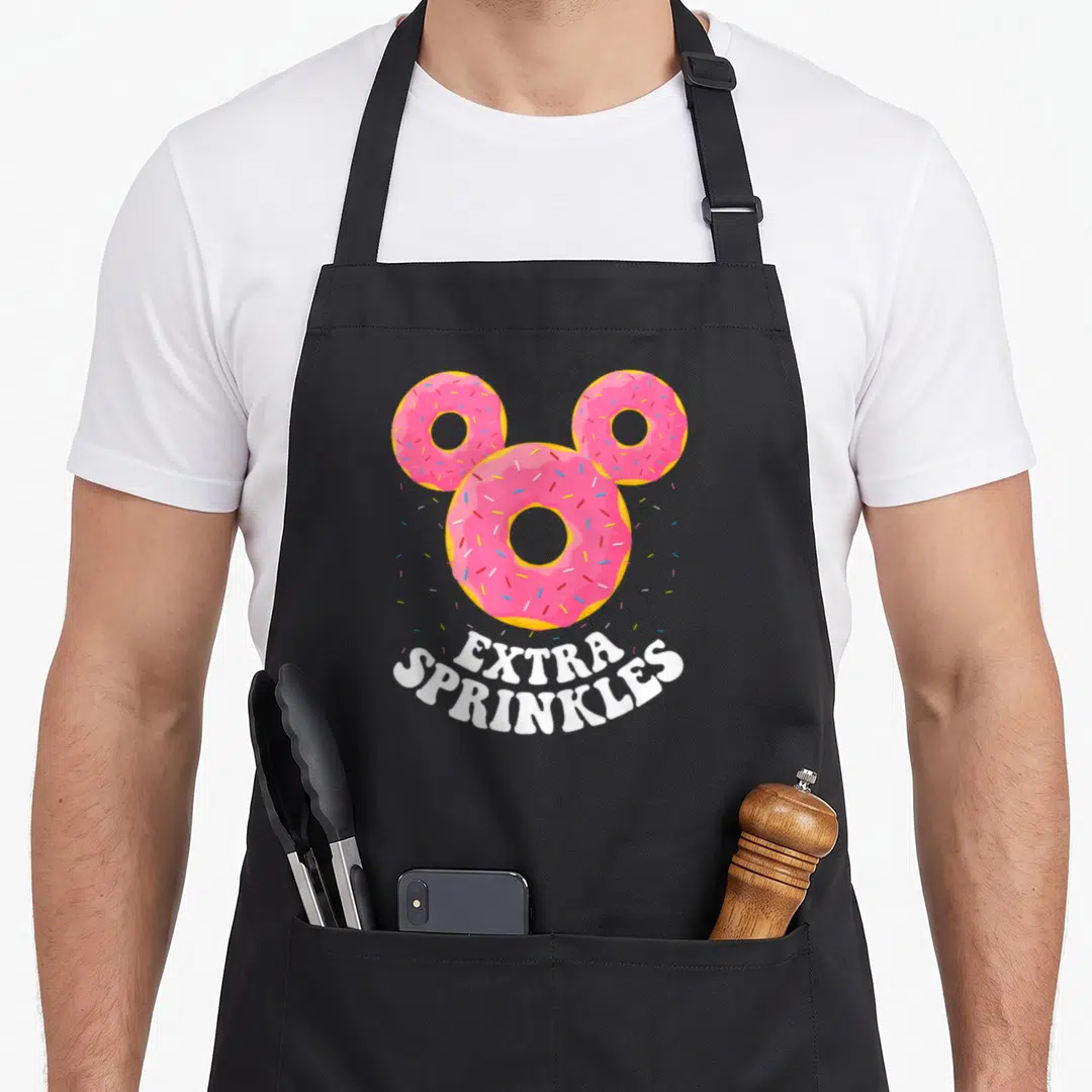 Ear sprinkle apron - Three pink donuts in mouse ear shape with white EXTRA SPRINKLES text — shown on a black cotton canvas apron against a white background