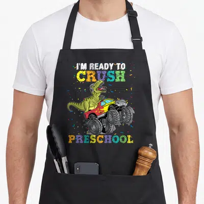 Rex truck apron - Cartoon dinosaur in monster truck with I'm Ready To Crush Preschool text and confetti — shown on a black cotton canvas apron against a white background