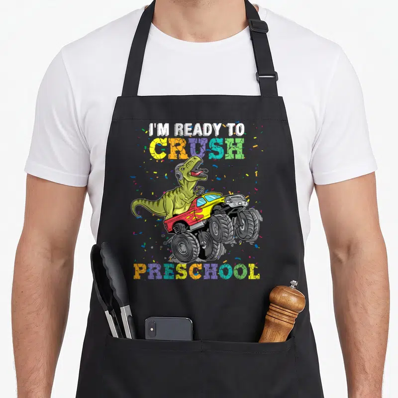 Rex truck apron - Cartoon dinosaur in monster truck with I'm Ready To Crush Preschool text and confetti — shown on a black cotton canvas apron against a white background