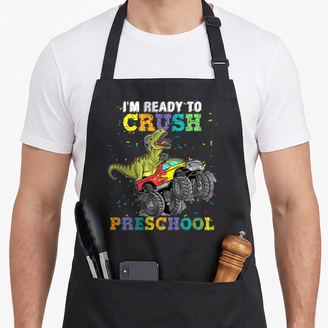 Rex truck apron - Cartoon dinosaur in monster truck with I'm Ready To Crush Preschool text and confetti — shown on a black cotton canvas apron against a white background