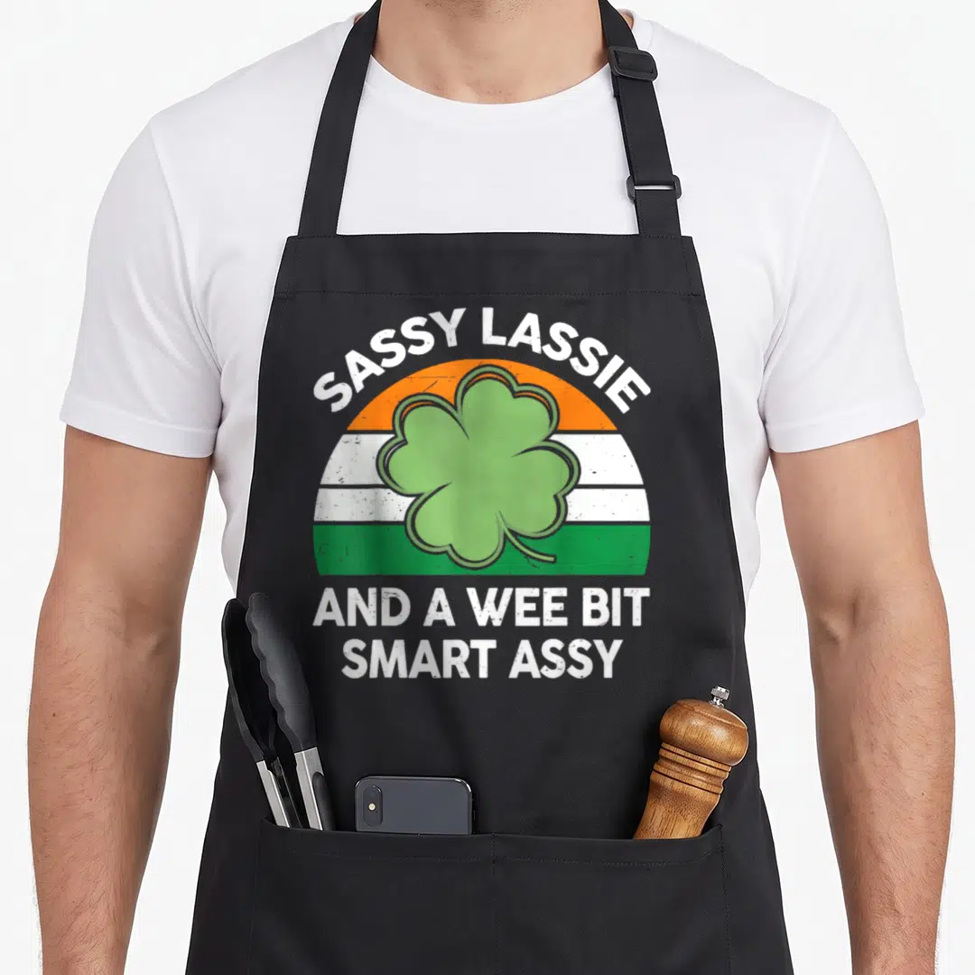 Sassy irish aprons - SASSY LASSIE text over four-leaf clover on orange white green distressed arc — shown on a black cotton canvas apron against a white background