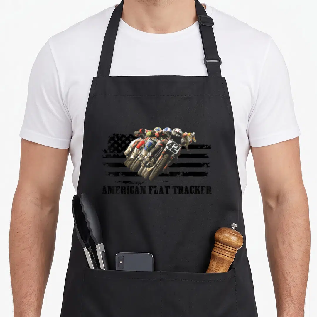 Flat racing sports apron - Motorcyclists racing in front of distressed black USA flag with AMERICAN FLAT TRACKER text — shown on a black cotton canvas apron against a white background
