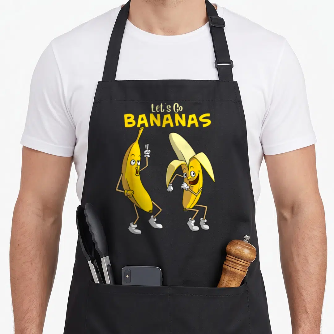 Bananas fruit apron - Two dancing cartoon bananas under Let's Go BANANAS text in yellow white tan — shown on a black cotton canvas apron against a white background