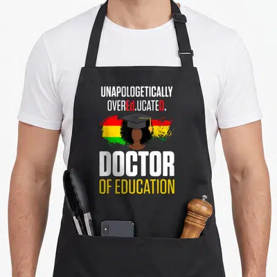 Education doctor apron - Unapologetically Overeducated text with woman in graduation cap and brush strokes — shown on a black cotton canvas apron against a white background