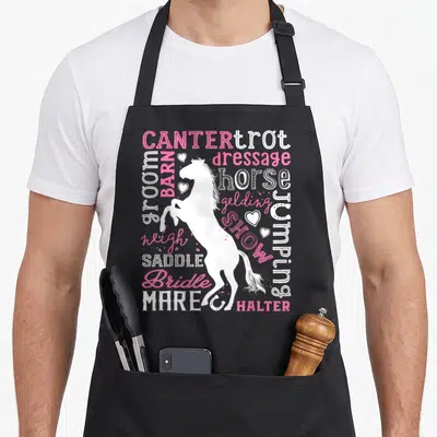 Typography horse apron - White rearing horse silhouette surrounded by pink and white equestrian text terms and small — shown on a black cotton canvas apron against a white background