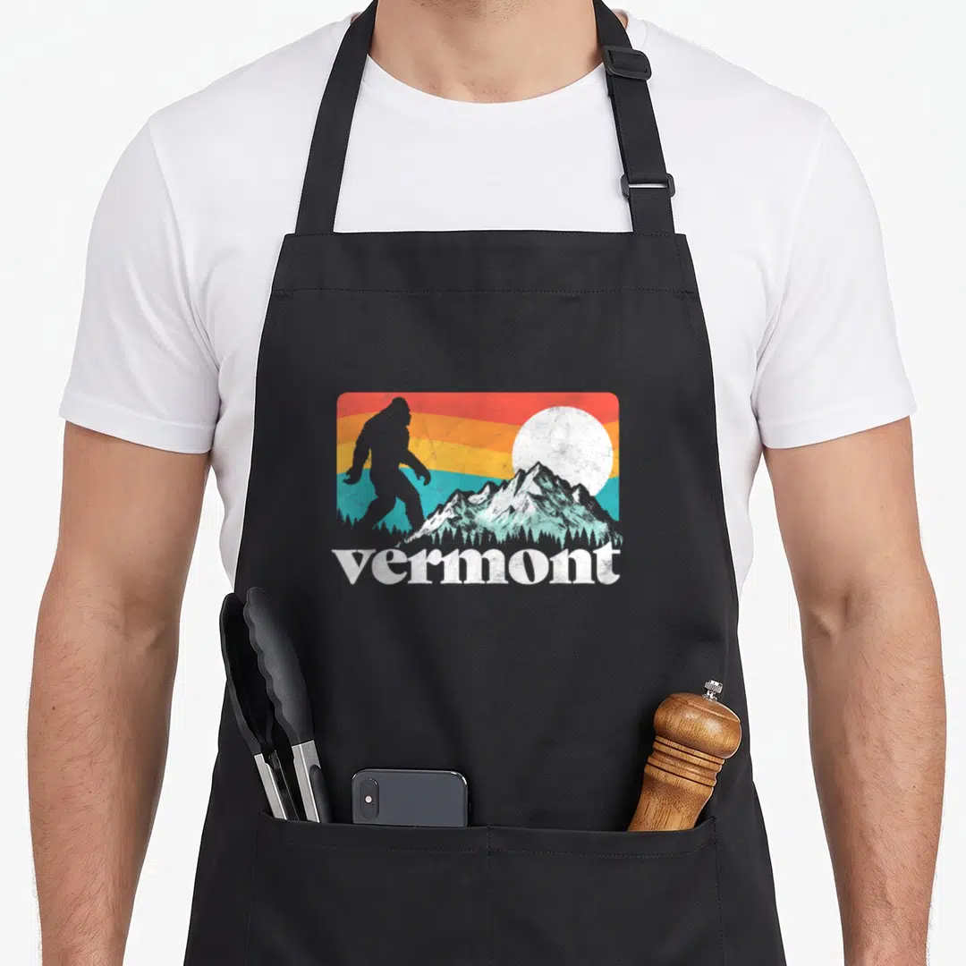 Pride vermont apron - Bigfoot silhouette on red orange yellow teal stripes with white mountains and vermont text — shown on a black cotton canvas apron against a white background