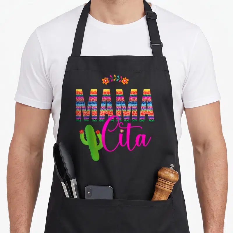 Mamacita cactus apron - Colorful MAMA cita text with striped patterns, floral arch, and green cactus illustration — shown on a black cotton canvas apron against a white background