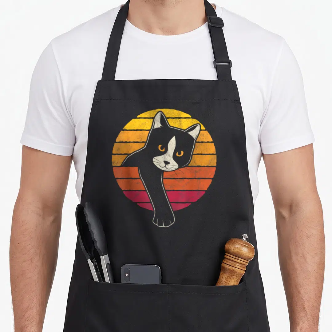 Sunset tuxedo apron - Grey bicolor cat silhouette over yellow orange red striped circle — shown on a black cotton canvas apron against a white background