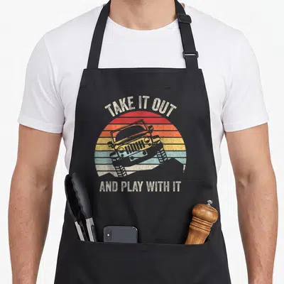 Catch fried apron - Distressed off-road vehicle on striped red orange yellow blue sun with arched text — shown on a black cotton canvas apron against a white background