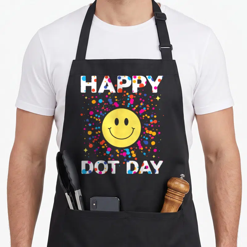 Colorful polka dot apron - HAPPY DOT DAY text with yellow smiley face and multicolored dots — shown on a black cotton canvas apron against a white background