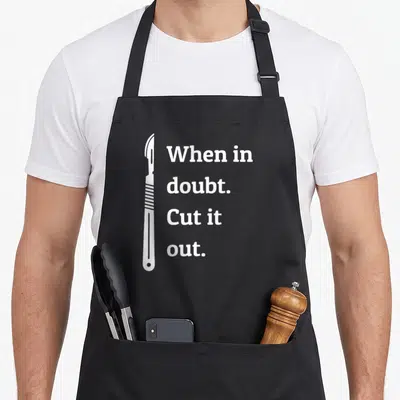 Doubt surgeon apron - White scalpel illustration beside stacked text When in doubt Cut it out — shown on a black cotton canvas apron against a white background