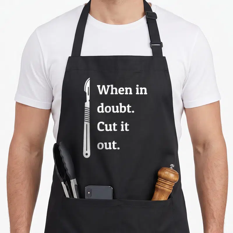 Doubt surgeon apron - White scalpel illustration beside stacked text When in doubt Cut it out — shown on a black cotton canvas apron against a white background