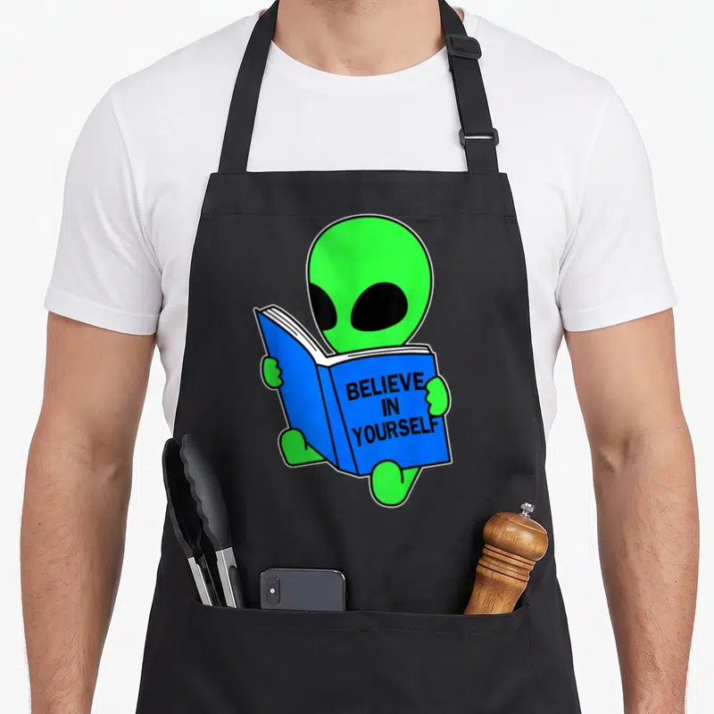 Yourself alien ufo apron - Green alien holding open blue book with believe in yourself text — shown on a black cotton canvas apron against a white background