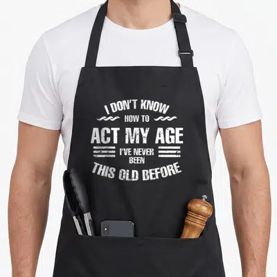 Cute act age apron - White distressed text reading I dont know how to act my age with swirls and horizontal lines — shown on a black cotton canvas apron against a white background