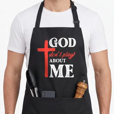 Play faith christian apron - Red brush-stroke cross beside white distressed text reading GOD don't play ABOUT ME — shown on a black cotton canvas apron against a white background