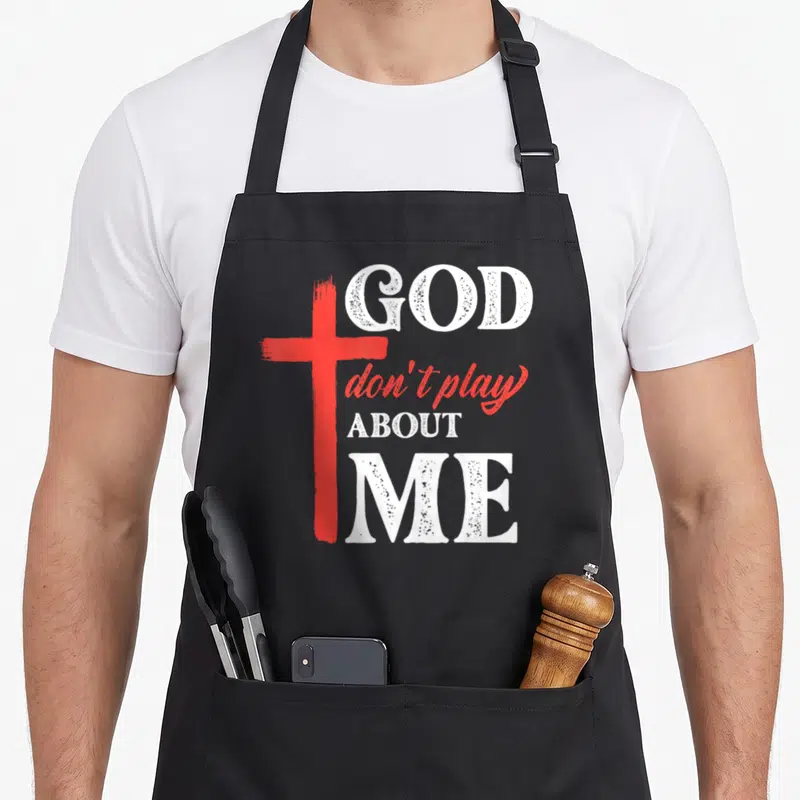 Play faith christian apron - Red brush-stroke cross beside white distressed text reading GOD don't play ABOUT ME — shown on a black cotton canvas apron against a white background