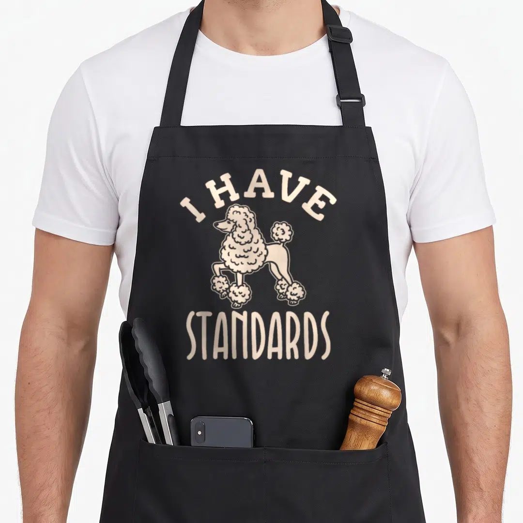 Breed standard size of apron - Beige cartoon poodle standing between arched I HAVE and straight STANDARDS text — shown on a black cotton canvas apron against a white background