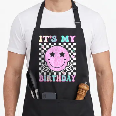 Groovy custom face apron - Pink leopard smiley face with star eyes and peace hands over checkerboard with IT'S MY — shown on a black cotton canvas apron against a white background