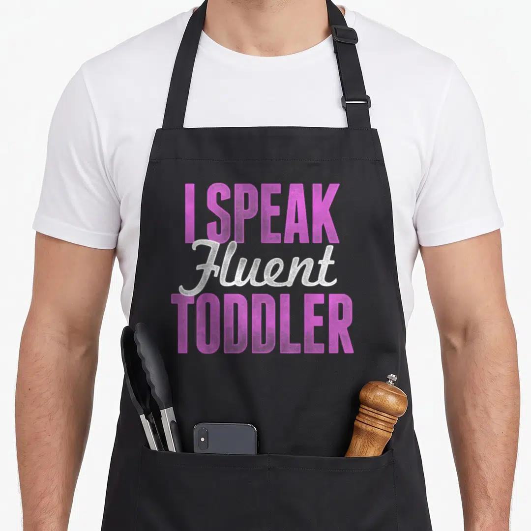 Speak toddler mom apron - Pink and white stacked text reading I SPEAK Fluent TODDLER — shown on a black cotton canvas apron against a white background