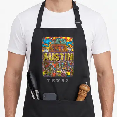 Live texas aprons - Cartoon AUSTIN letters in yellow with patterns above a colorful crowd of people — shown on a black cotton canvas apron against a white background