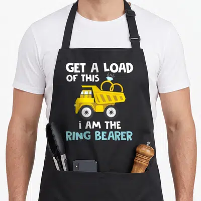 Wedding truck apron - Cartoon yellow dump truck carrying two gold rings with diamond tops and bold white text — shown on a black cotton canvas apron against a white background