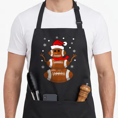 Football snowman apron - Three brown footballs stacked as a snowman wearing a red Santa hat and scarf with white — shown on a black cotton canvas apron against a white background