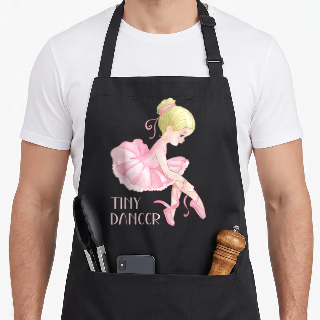 Tiny ballerina apron - Cartoon blonde girl in pink tutu tying ballet shoes above stacked TINY DANCER text — shown on a black cotton canvas apron against a white background