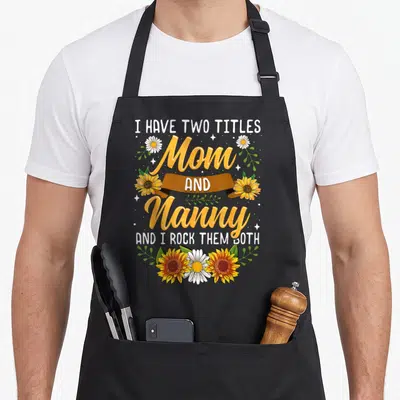 Title nanny apron - Stacked typography Mom Nanny with sunflowers daisies brown ribbon white sparkles on black background — shown on a black cotton canvas apron against a white background