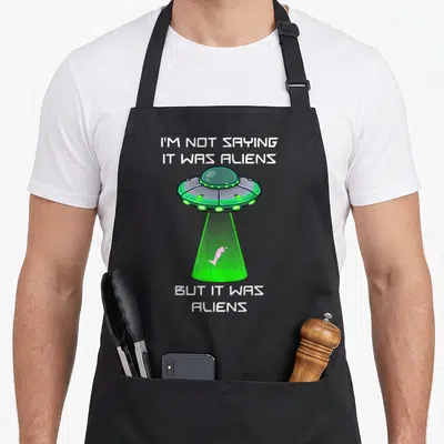 Foot leg apron - Cartoon UFO lifting pink leg in green beam with sarcastic white text on grey background — shown on a black cotton canvas apron against a white background