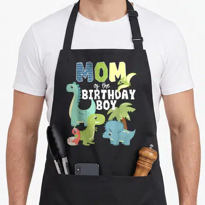 Theme dinosaur apron - Cartoon dinos and palm tree surround MOM of the BIRTHDAY BOY text in teal green and blue — shown on a black cotton canvas apron against a white background