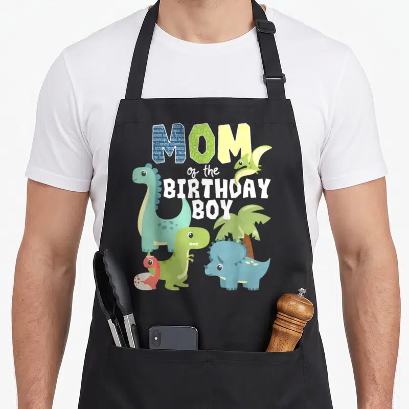 Theme dinosaur apron - Cartoon dinos and palm tree surround MOM of the BIRTHDAY BOY text in teal green and blue — shown on a black cotton canvas apron against a white background