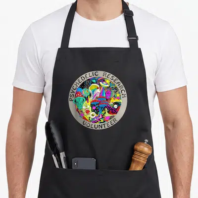 Psychedelic mushroom apron - Circular PSYCHEDELIC RESEARCH VOLUNTEER text bordering a multicolored cartoon collage — shown on a black cotton canvas apron against a white background