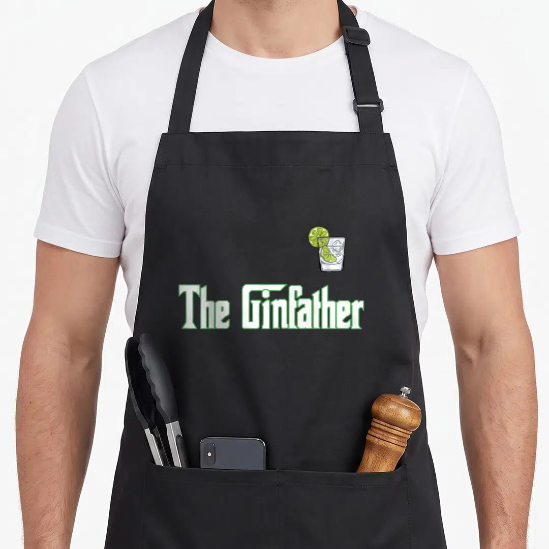 Ginfather gin apron - The Ginfather text with cartoon gin and tonic glass and lime slice — shown on a black cotton canvas apron against a white background