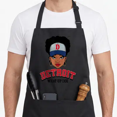 Melanin detroit apron - Cartoon woman wearing a cap above arched red DETROIT text and straight black WHAT UP DOE — shown on a black cotton canvas apron against a white background