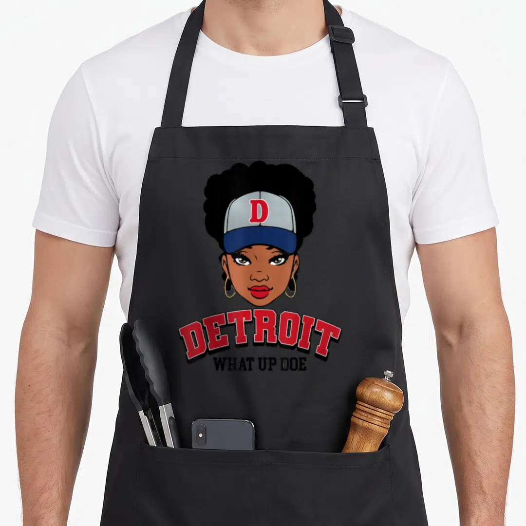 Melanin detroit apron - Cartoon woman wearing a cap above arched red DETROIT text and straight black WHAT UP DOE — shown on a black cotton canvas apron against a white background