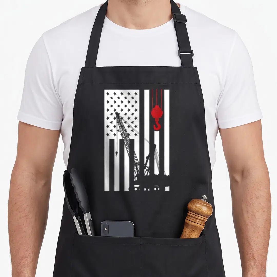 Crane us aprons - Grey crane silhouette over white stripes and grey stars with hanging red hook — shown on a black cotton canvas apron against a white background