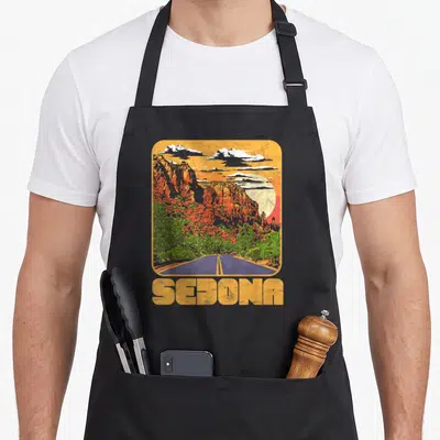 Sedona road apron - Retro landscape with rounded corners, orange cliffs, blue road, and yellow SEDONA text — shown on a black cotton canvas apron against a white background