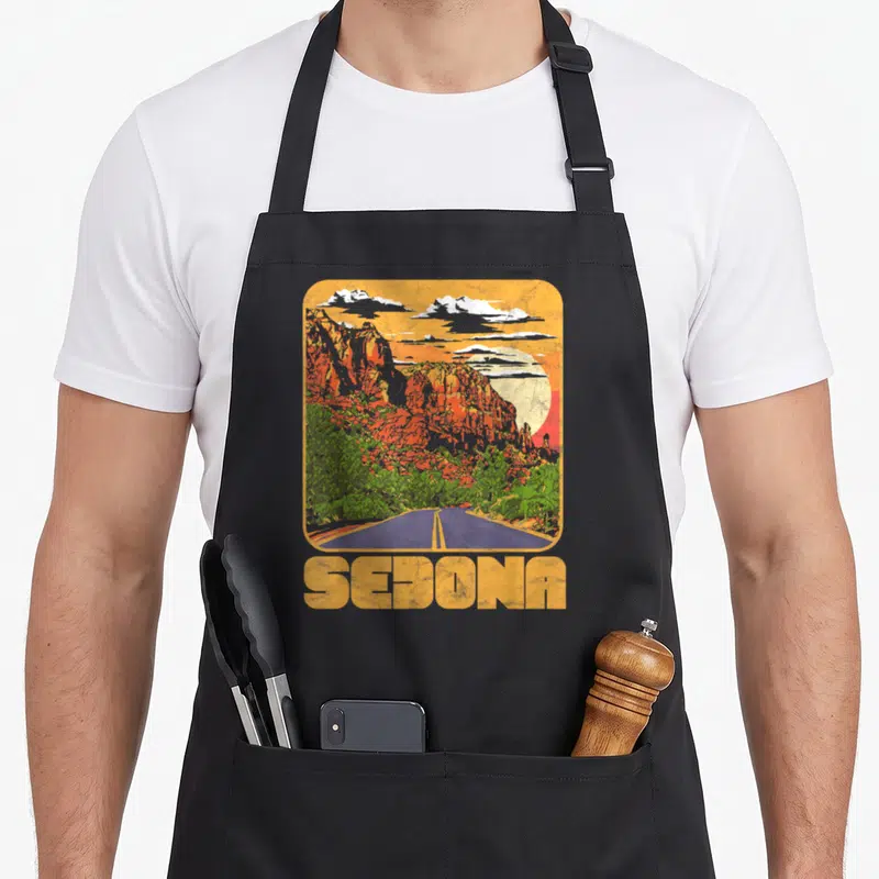 Sedona road apron - Retro landscape with rounded corners, orange cliffs, blue road, and yellow SEDONA text — shown on a black cotton canvas apron against a white background