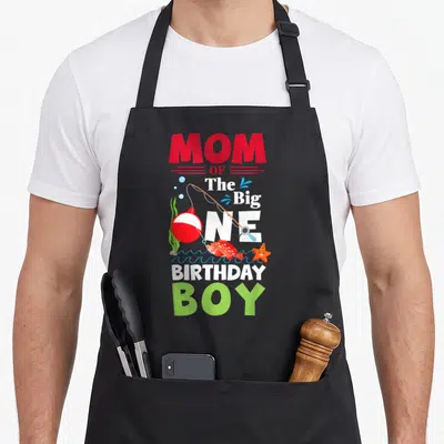 Birthday fishing fish apron - Mom of the Big One Birthday Boy text with cartoon fish, rod, starfish, and waves — shown on a black cotton canvas apron against a white background