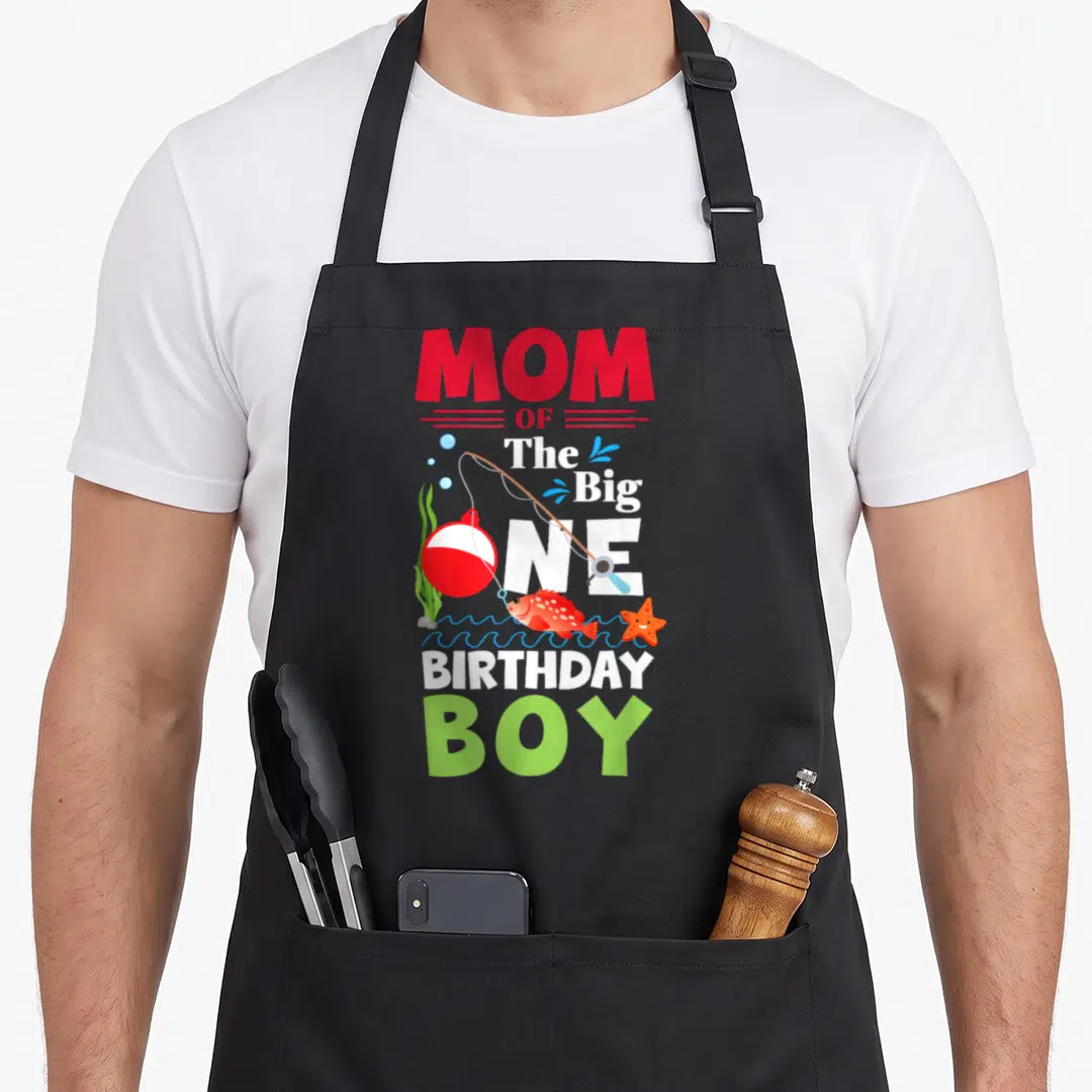 Birthday fishing fish apron - Mom of the Big One Birthday Boy text with cartoon fish, rod, starfish, and waves — shown on a black cotton canvas apron against a white background