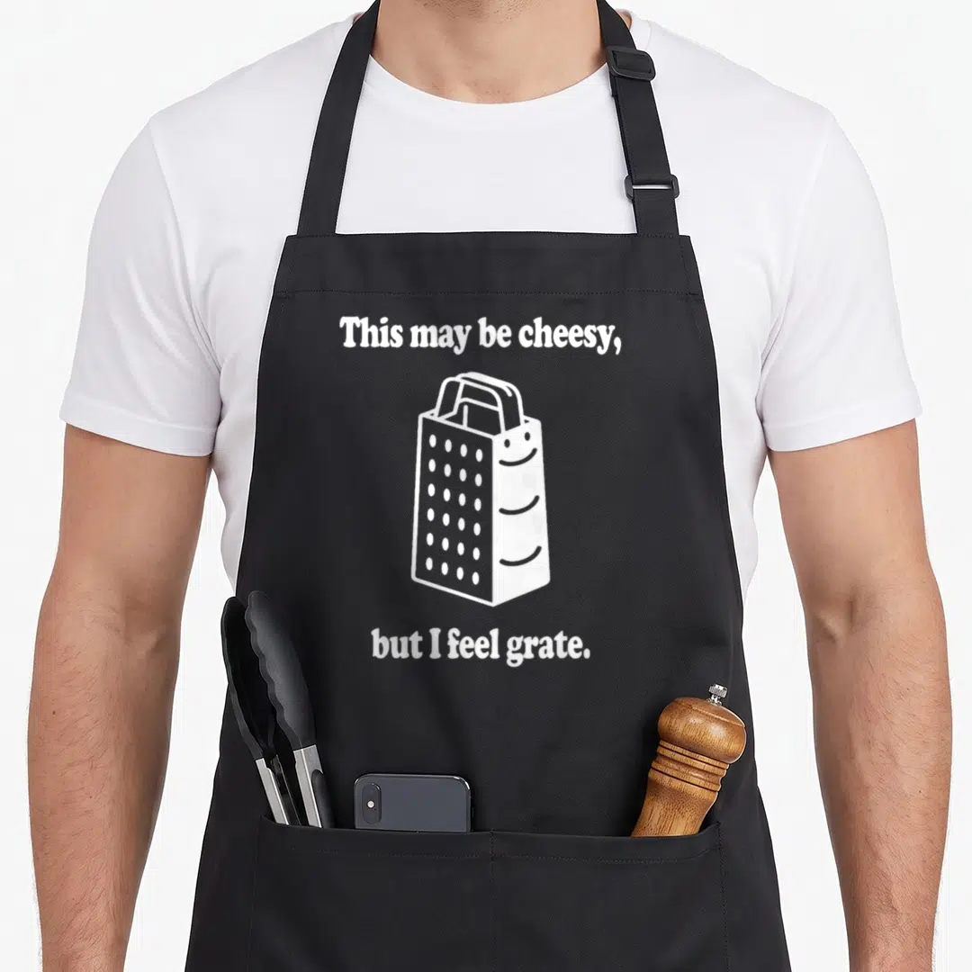 May cheesy aprons - White cartoon cheese grater with smiley face between two lines of text reading This may be cheesy — shown on a black cotton canvas apron against a white background