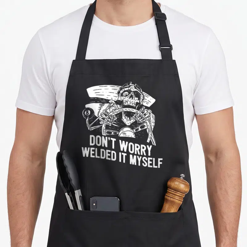 Welder builder apron - White skeleton drawing gripping handlebars above stacked text DON'T WORRY WELDED IT MYSELF — shown on a black cotton canvas apron against a white background