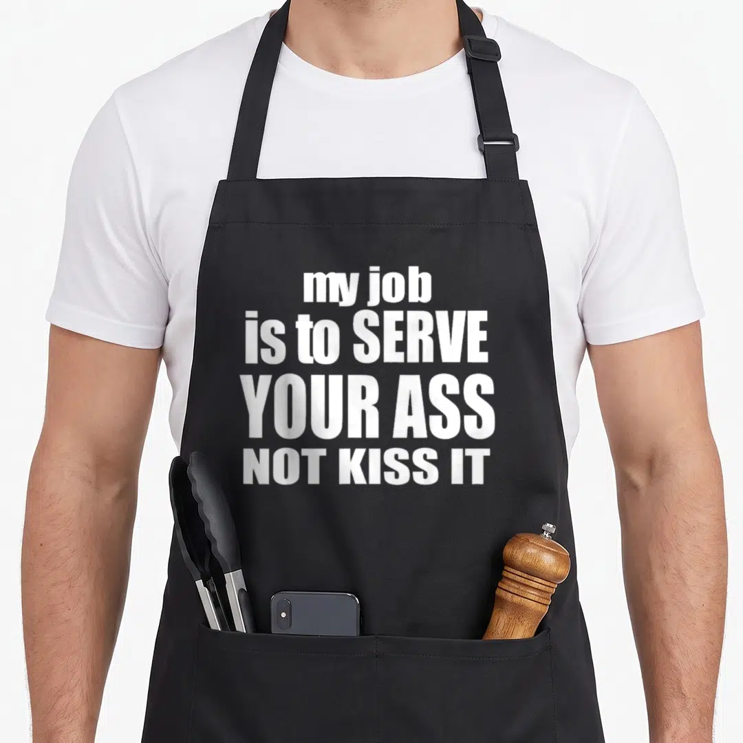 Kiss to serve man apron - White stacked text reading my job is to serve your ass not kiss it on black fabric — shown on a black cotton canvas apron against a white background